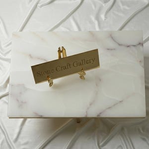 Könnte beinhalten: Eine rechteckige, weiße Marmorplatte mit dezenten rosa Adern. Ein goldfarbenes Schild mit der Aufschrift "Stone Craft Gallery" ist mit einem goldfarbenen Ständer am Marmor befestigt. Der Hintergrund ist ein weißer, seidiger Stoff.