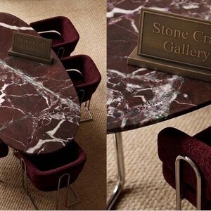 May include: A dining table with a red marble top and white veining, surrounded by burgundy chairs with silver legs. A bronze sign on the table reads "Stone Craft Gallery". The table is oval-shaped and the chairs have a curved design.