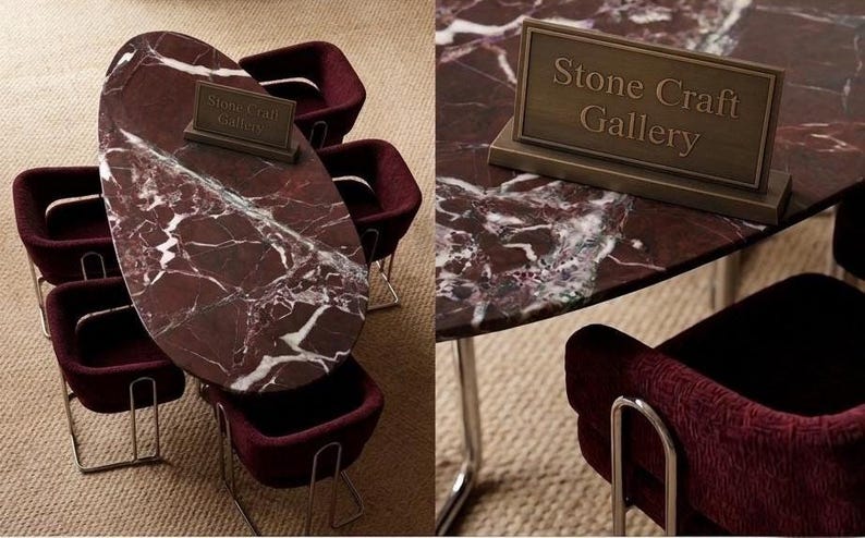May include: A dining table with a red marble top and white veining, surrounded by burgundy chairs with silver legs. A bronze sign on the table reads "Stone Craft Gallery". The table is oval-shaped and the chairs have a curved design.