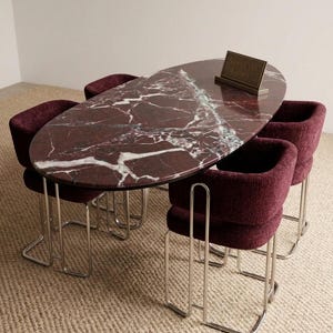 May include: An oval dining table with a burgundy marble top and a white veined pattern. The table is surrounded by four burgundy upholstered chairs with chrome legs. A small sign on the table reads "Stone Craft Gallery."