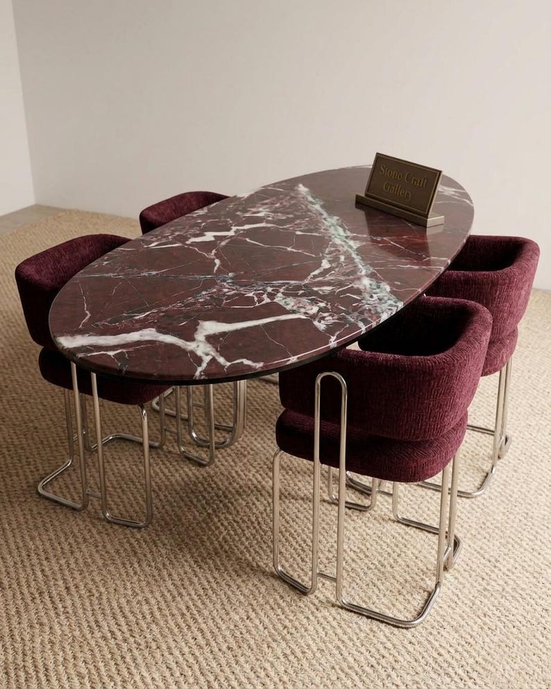May include: An oval dining table with a burgundy marble top and a white veined pattern. The table is surrounded by four burgundy upholstered chairs with chrome legs. A small sign on the table reads "Stone Craft Gallery."