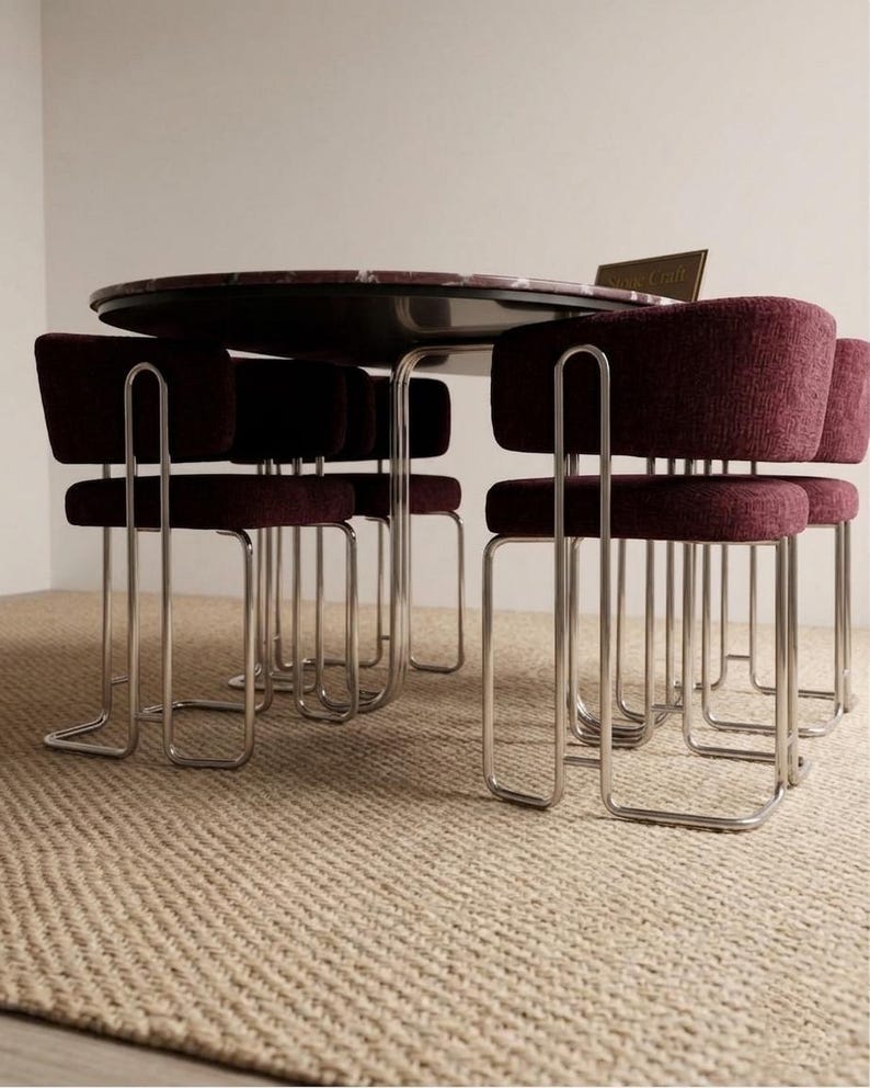 May include: A round dining table with a dark marble top is surrounded by six burgundy upholstered chairs with chrome frames. The chairs have a unique, curved design. The table and chairs are set on a beige woven rug.