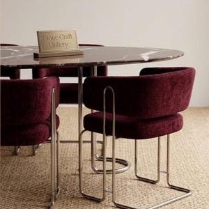May include: A modern dining set featuring a round marble-topped table and four burgundy upholstered chairs with chrome legs. The table has a sign that reads "Stone Craft Gallery". The chairs have a curved back design.