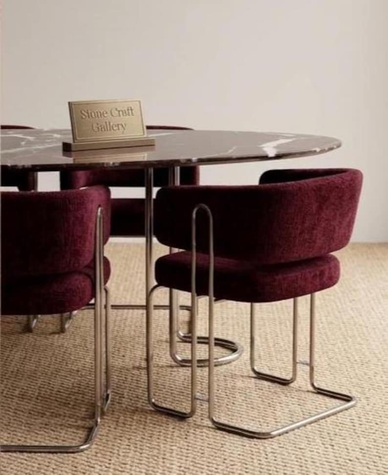 May include: A modern dining set featuring a round marble-topped table and four burgundy upholstered chairs with chrome legs. The table has a sign that reads "Stone Craft Gallery". The chairs have a curved back design.