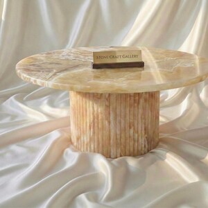 May include: A round, beige onyx coffee table with a cylindrical base. The table has a smooth, polished surface and a natural, veined pattern. The base features vertical, textured lines. A small sign that reads "STONE CRAFT GALLERY" rests on the table.