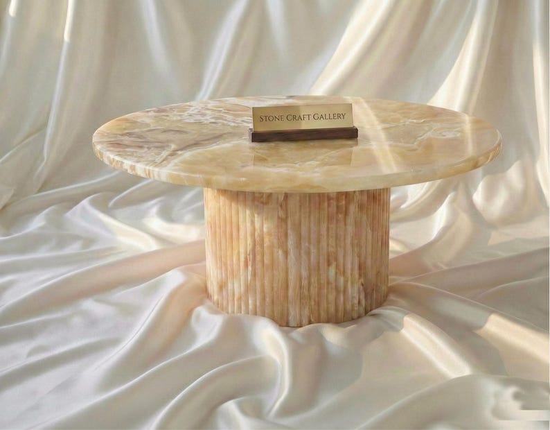 May include: A round, beige onyx coffee table with a cylindrical base. The table has a smooth, polished surface and a natural, veined pattern. The base features vertical, textured lines. A small sign that reads "STONE CRAFT GALLERY" rests on the table.
