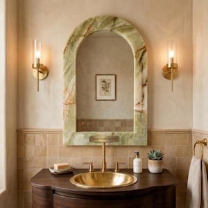 May include: An arched mirror with a green and white veined stone frame featuring the text Stone Craft Gallery, positioned above a dark wood vanity with a gold sink, gold faucet, two gold wall sconces, a soap dish, and a small succulent.