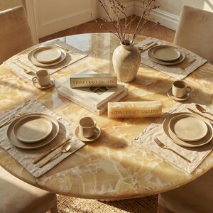 May include: A round dining table with a marble-like surface is set for a meal. Each place setting includes a plate, bowl, cup, and gold-colored silverware on a linen placemat. A vase with branches and books are in the center. The text "STONE CRAFT GALLERY" is visible.