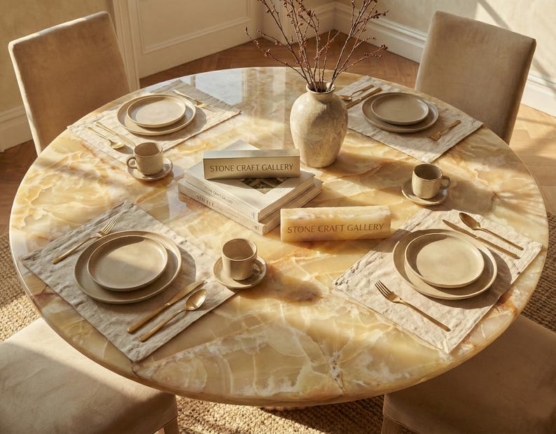 May include: A round dining table with a marble-like surface is set for a meal. Each place setting includes a plate, bowl, cup, and gold-colored silverware on a linen placemat. A vase with branches and books are in the center. The text "STONE CRAFT GALLERY" is visible.