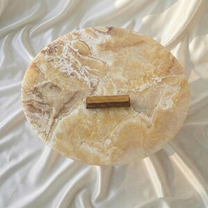 May include: A round, marble-like stone serving board with a warm, beige and brown color palette. The board has a natural, veined pattern and a small wooden label that reads "STONE CRAFT GALLERY".