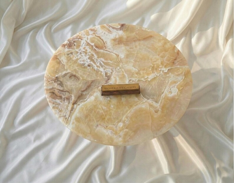 May include: A round, marble-like stone serving board with a warm, beige and brown color palette. The board has a natural, veined pattern and a small wooden label that reads "STONE CRAFT GALLERY".