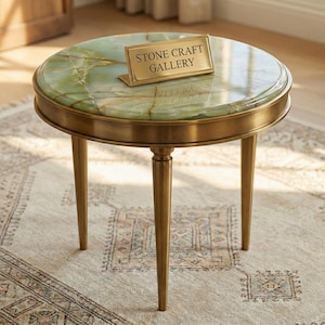 May include: A round side table with a green marble top and brass-colored metal frame. The table has three slender legs and a small rectangular sign that reads "STONE CRAFT GALLERY". The table is on a patterned rug.