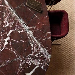 May include: Oval-shaped marble table with a deep red and white veined pattern. The table has a bronze sign that reads "Stone Craft Gallery". Two burgundy chairs are visible in the background.