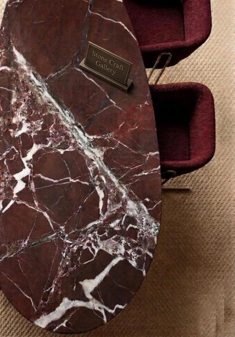 May include: Oval-shaped marble table with a deep red and white veined pattern. The table has a bronze sign that reads "Stone Craft Gallery". Two burgundy chairs are visible in the background.