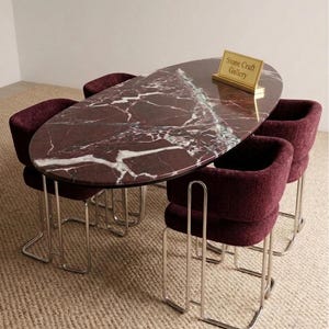 May include: An oval dining table with a burgundy marble top and four matching burgundy upholstered chairs. The chairs have chrome legs. A small gold sign on the table reads "Stone Craft Gallery."