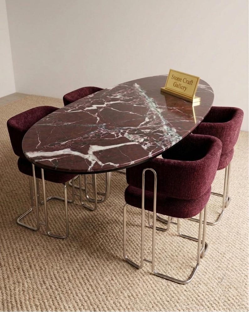 May include: An oval dining table with a burgundy marble top and four matching burgundy upholstered chairs. The chairs have chrome legs. A small gold sign on the table reads "Stone Craft Gallery."