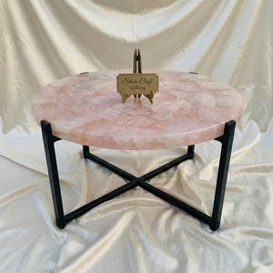 May include: A circular coffee table featuring a pink quartz surface and black metal legs. The table has a distinctive, natural stone aesthetic. A small sign reading "Stone Craft Gallery" is displayed on the table.