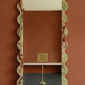 May include: A tall rectangular mirror featuring a wavy, scalloped frame made of light green stone with brown veining. The mirror reflects a small gold-colored sign on a thin stand that reads Stone Craft Gallery in black text.