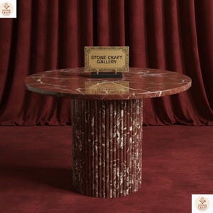 May include: A round, red marble table with white veining. The table has a cylindrical base with vertical, textured lines. A gold sign with the text "STONE CRAFT GALLERY" sits on top of the table.