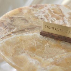 May include: A close-up of a round, polished stone table with a natural, marbled pattern in shades of beige and brown. A brass sign with the text "STONE CRAFT GALLERY" rests on the table's edge.