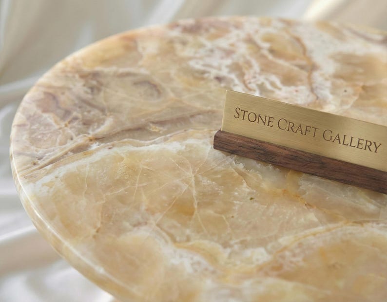 May include: A close-up of a round, polished stone table with a natural, marbled pattern in shades of beige and brown. A brass sign with the text "STONE CRAFT GALLERY" rests on the table's edge.