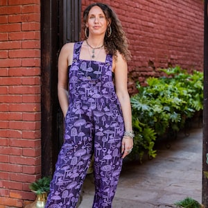 May include: Purple overalls with a mushroom print. The overalls have adjustable straps and a patch pocket on the chest.