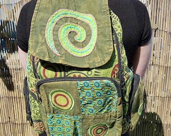 May include: Green backpack with a patchwork design featuring circles and geometric patterns. The flap has a stitched spiral design in shades of green, pink, and blue. The backpack has multiple pockets and adjustable straps.