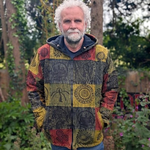 Può includere: Giacca con cappuccio e zip in patchwork, con quadrati rossi, gialli e neri, ognuno con un motivo unico. La giacca è indossata sopra una maglietta blu scuro e abbinata a jeans blu. Lo sfondo comprende alberi e vegetazione.
