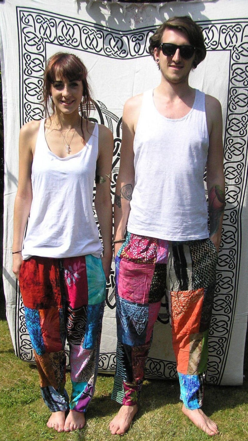 May include: Two people wearing white tank tops and colorful patchwork pants stand in front of a white tapestry with a black Celtic knot design.