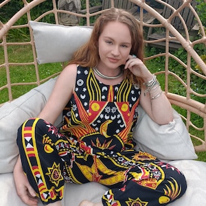 Puede incluir: Una mujer lleva un mono negro y amarillo con estampado celestial. El mono tiene un top sin mangas y pantalones anchos. La mujer está sentada en una silla de mimbre.