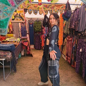 May include: A woman wearing a black crop top with white embroidery and black pants with white geometric patterns. She is standing in a market stall with colorful fabrics and clothing hanging from the ceiling.