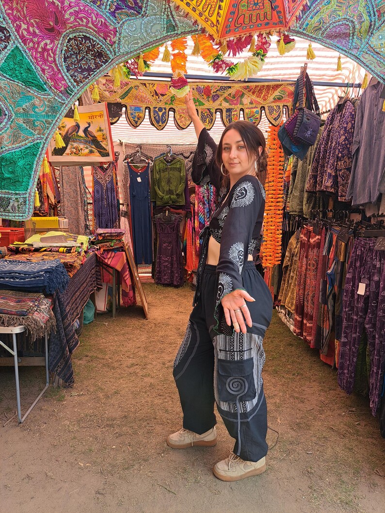 May include: A woman wearing a black crop top with white embroidery and black pants with white geometric patterns. She is standing in a market stall with colorful fabrics and clothing hanging from the ceiling.