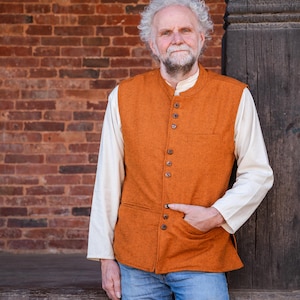 Op de afbeelding: Een oranje vest met een opstaande kraag en knoopsluiting. Het vest wordt gedragen over een crèmekleurig shirt met lange mouwen en gecombineerd met een blauwe spijkerbroek. De achtergrond toont een bakstenen muur en een houten paal.