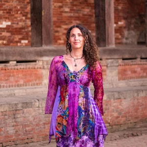 May include: A woman wearing a purple and blue patterned dress with long sleeves. The dress has a tie at the neckline and a layered skirt. She is standing in front of a brick wall.