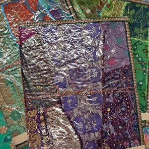 May include: A collection of handmade paper notebooks with patchwork designs. The notebooks feature a variety of fabrics in shades of purple, green, and gold, with detailed embroidery and embellishments. Each notebook is wrapped in clear plastic.
