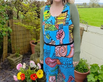 May include: A woman wearing a blue jumpsuit with a colorful abstract pattern. The jumpsuit has a black belt and a scoop neck. The woman is standing in a garden with flowers and plants.