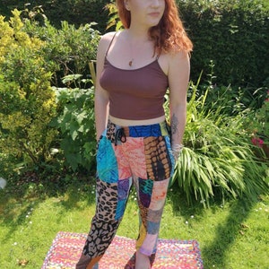 May include: A woman wearing a brown tank top and colorful patchwork pants. She is standing on a multi-colored rug in a grassy area.