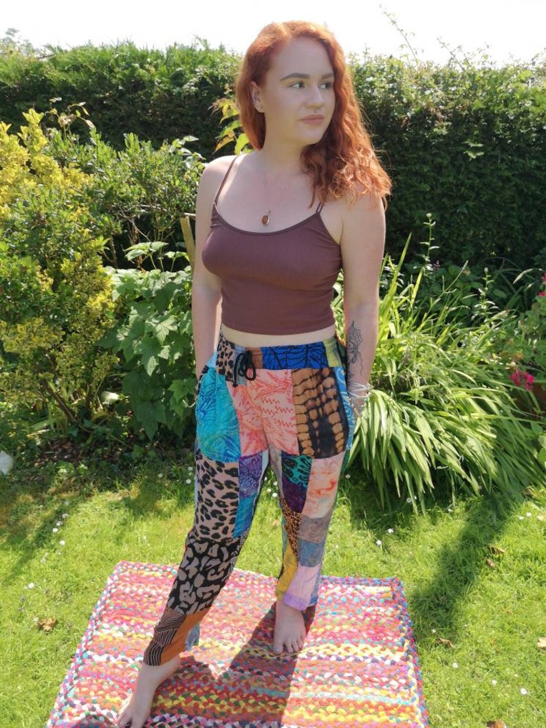 May include: A woman wearing a brown tank top and colorful patchwork pants. She is standing on a multi-colored rug in a grassy area.
