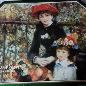 May include: A vintage tin box featuring a painting of two women in a garden setting. The woman in the foreground is wearing a red hat and a black dress. The woman in the background is wearing a blue and white hat and a white dress. The text "Russell Stover Candies" is printed on the box.