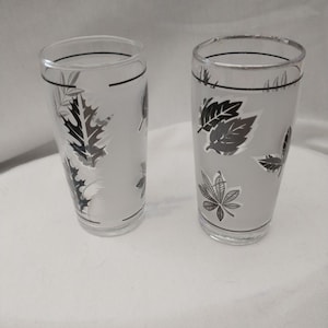 May include: Two clear glass tumblers with a silver and black leaf design. The glasses are frosted and have a black line around the top.
