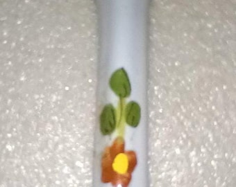 May include: A white ceramic spoon featuring a hand-painted floral design on the handle. The pattern consists of green leaves, a single orange flower with a yellow center, and a blue flower with a yellow center arranged in a vertical row.