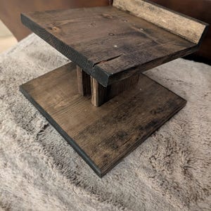 Op de afbeelding: Een donkerbruin gebeitste houten standaard met een platte rechthoekige voet, een centrale verticale steunpilaar en een verhoogd platform met een kleine achterrand. De houtnerf is over de gehele structuur zichtbaar.