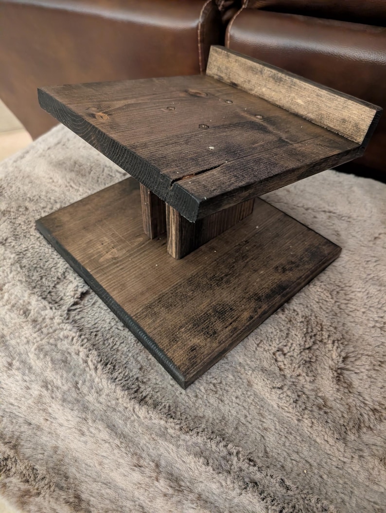 Op de afbeelding: Een donkerbruin gebeitste houten standaard met een platte rechthoekige voet, een centrale verticale steunpilaar en een verhoogd platform met een kleine achterrand. De houtnerf is over de gehele structuur zichtbaar.