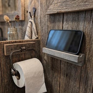 Könnte beinhalten: Ein rechteckiges graues Holzregal an einer Wand aus verwittertem Holz, auf dem ein schwarzes Smartphone in horizontaler Position steht. Unter dem Regal ist ein Metall-Toilettenpapierhalter mit einer Rolle weißem Toilettenpapier befestigt.