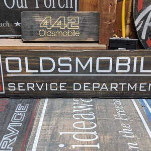 May include: A dark brown wooden sign with white lettering that reads "OLDSMOBILE SERVICE DEPARTMENT". The sign has a red and white logo on the left side.