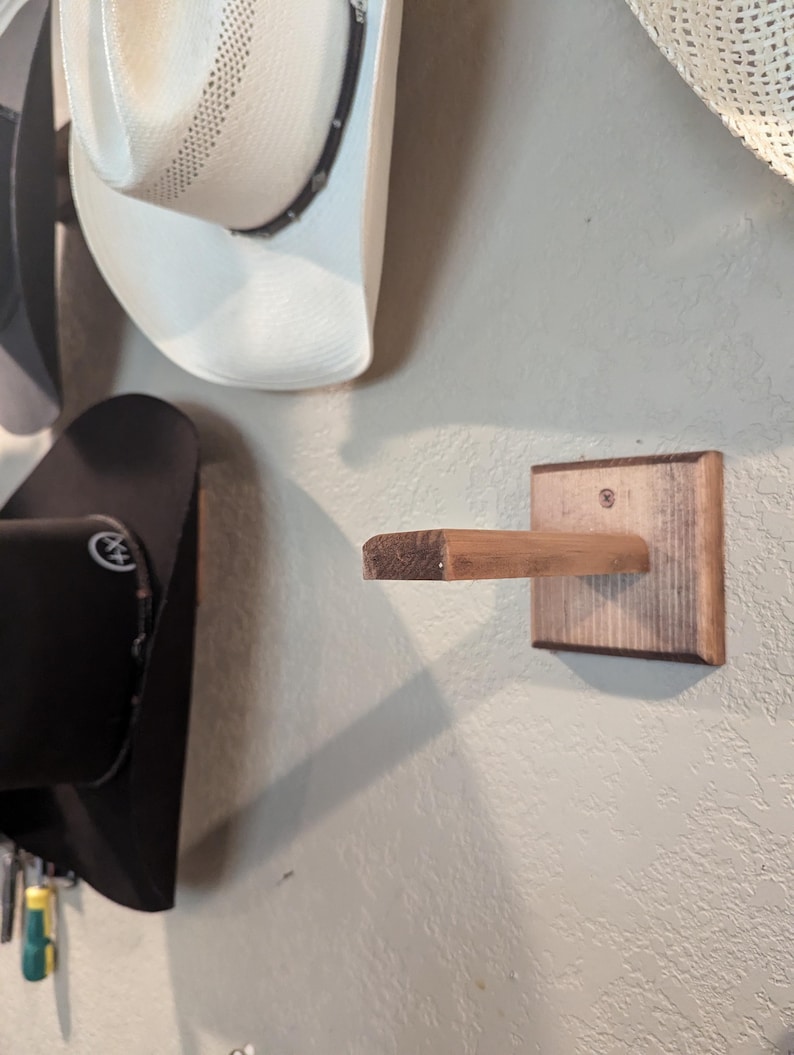 K&ouml;nnte beinhalten: Ein an einer Wand montierter Hutst&auml;nder aus Holz. Der St&auml;nder hat eine rechteckige Basis und einen horizontalen Arm, der nach au&szlig;en ragt. Der St&auml;nder ist aus hellem braunem Holz mit einer dunkleren braunen Beize an den Kanten.