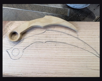 Puede incluir: Un cuchillo de madera de color claro estilo karambit con un anillo circular para el dedo en la base del mango, colocado directamente sobre un contorno a juego de la misma forma de cuchillo dibujado a lápiz en una tabla de madera clara.