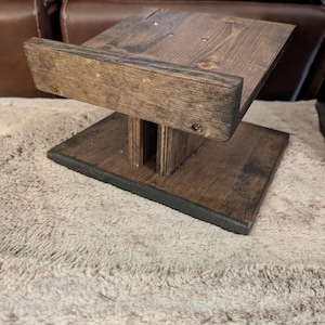 Op de afbeelding: Een donkerbruin gebeitste houten standaard met een rechthoekige voet, een centrale verticale steun en een schuin geplaatst bovenplatform met een opstaande rand aan de voorzijde. De houtnerf is zichtbaar over de gehele constructie met schroeven.