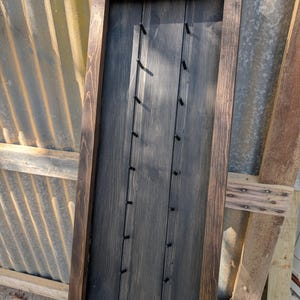 May include: A dark stained wooden display board with a rectangular frame. The board has vertical grooves and small pegs for hanging items. The frame is a darker brown, and the board itself is a charcoal gray color. The design is simple and rustic.