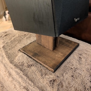 Op de afbeelding: Een zwarte luidspreker met een stoffen grille en een klein zilverkleurig logo staat op een donkerbruine houten standaard. De standaard heeft een vierkante voet en een verticaal steunstuk voor een stabiel platform voor de audioapparatuur.